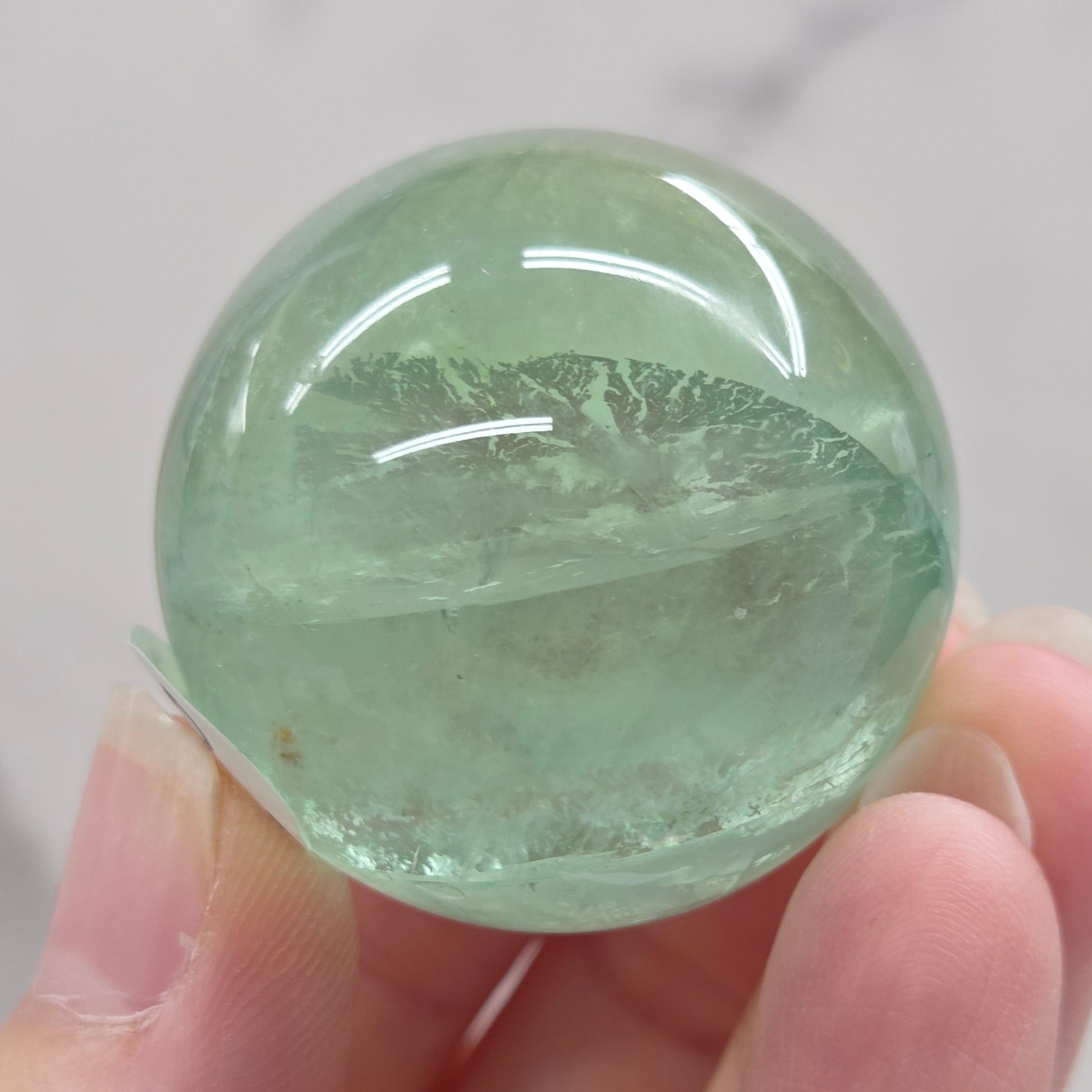 A close-up of a round, polished light green fluorite sphere mounted on a clear acrylic stand against a white marble-like background.