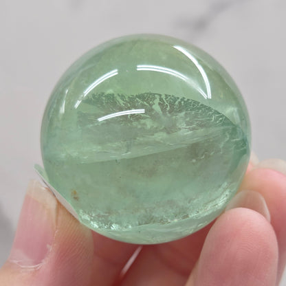 A close-up of a round, polished light green fluorite sphere mounted on a clear acrylic stand against a white marble-like background.