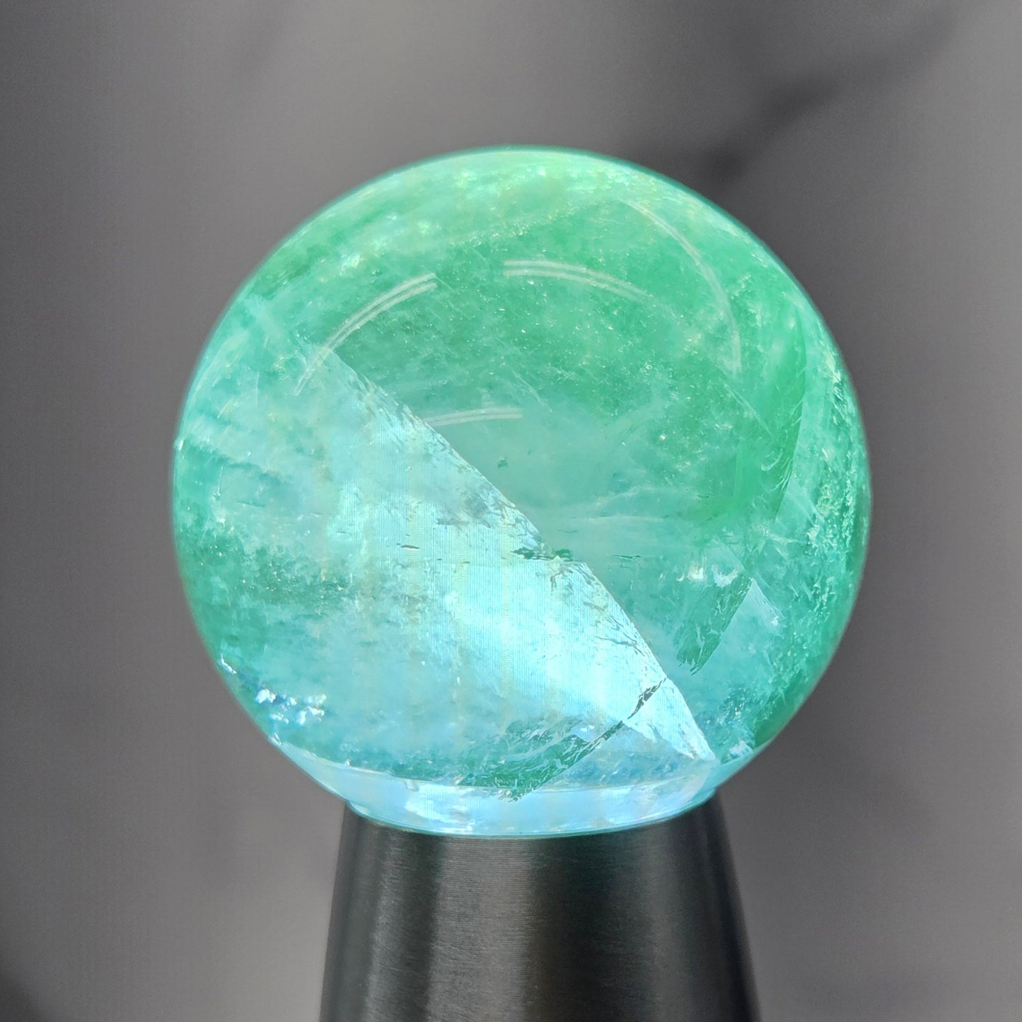 A hand holding a flashlight that illuminates a round, pale green translucent stone or crystal against a gray marble-like background.
