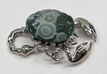 A silver crab-shaped brooch featuring a green and gray banded agate stone cabochon on its shell, set against a plain white background.