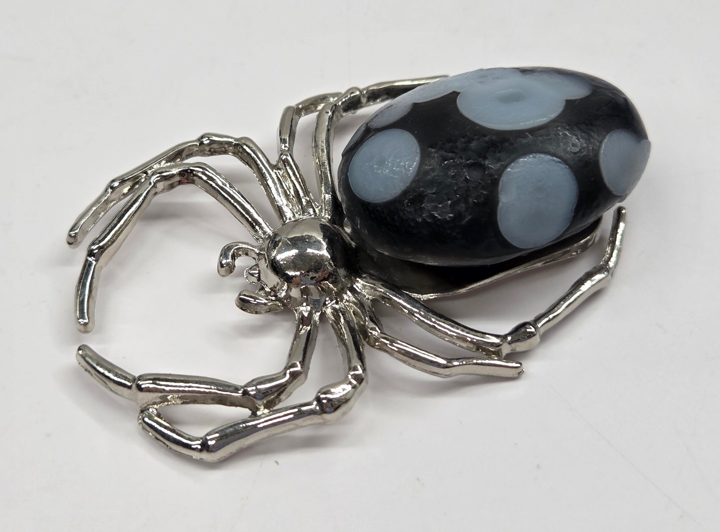 A silver-toned spider-shaped pendant or charm featuring a dark, marbled black and gray stone cabochon on its back, set against a plain light background.