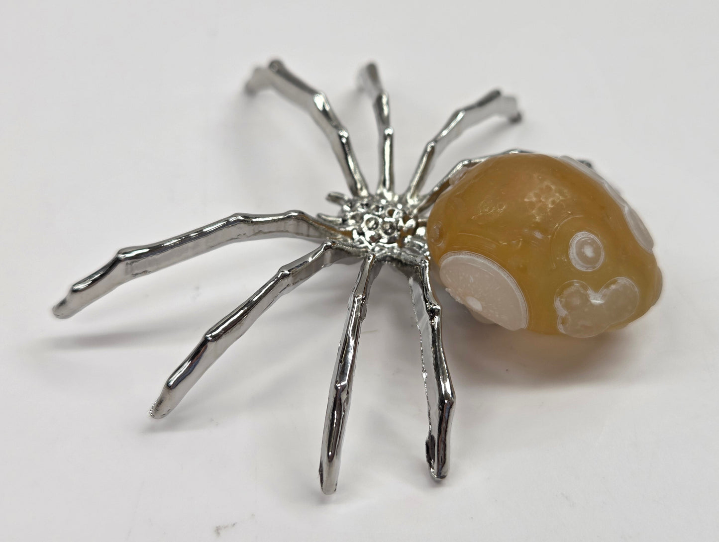 A spider-shaped pendant featuring a polished amber-brown bead body with white circular details and metallic silver legs, shown on a plain light background.