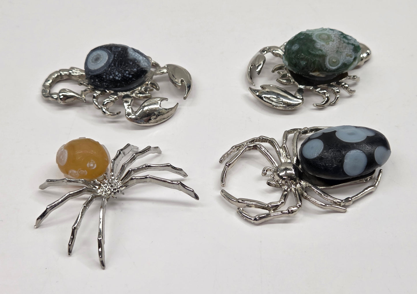 Four small spider and crab-shaped jewelry pieces with silver-toned metal bodies and decorative stone tops, arranged on a plain white background.