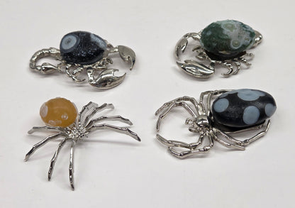 Four small spider and crab-shaped jewelry pieces with silver-toned metal bodies and decorative stone tops, arranged on a plain white background.