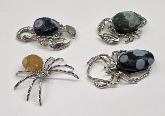Four small spider and crab-shaped jewelry pieces with silver-toned metal bodies and decorative stone tops, arranged on a plain white background.