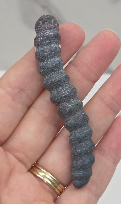 A close-up of a hand holding a dark gray, ribbed, worm-like textured object, with gold rings on one finger, against a light marble-patterned background.