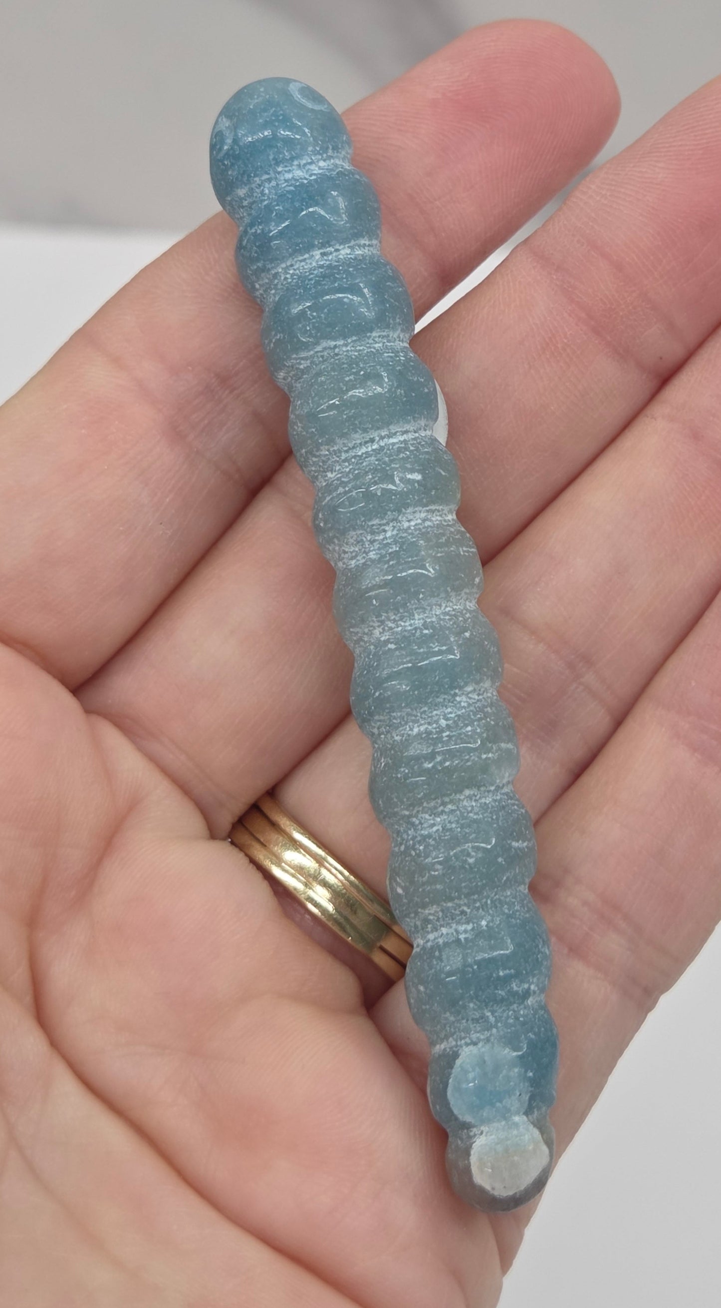A close-up of a hand holding a light blue, ribbed, worm-like textured object, with gold rings on one finger, against a light marble-patterned background.