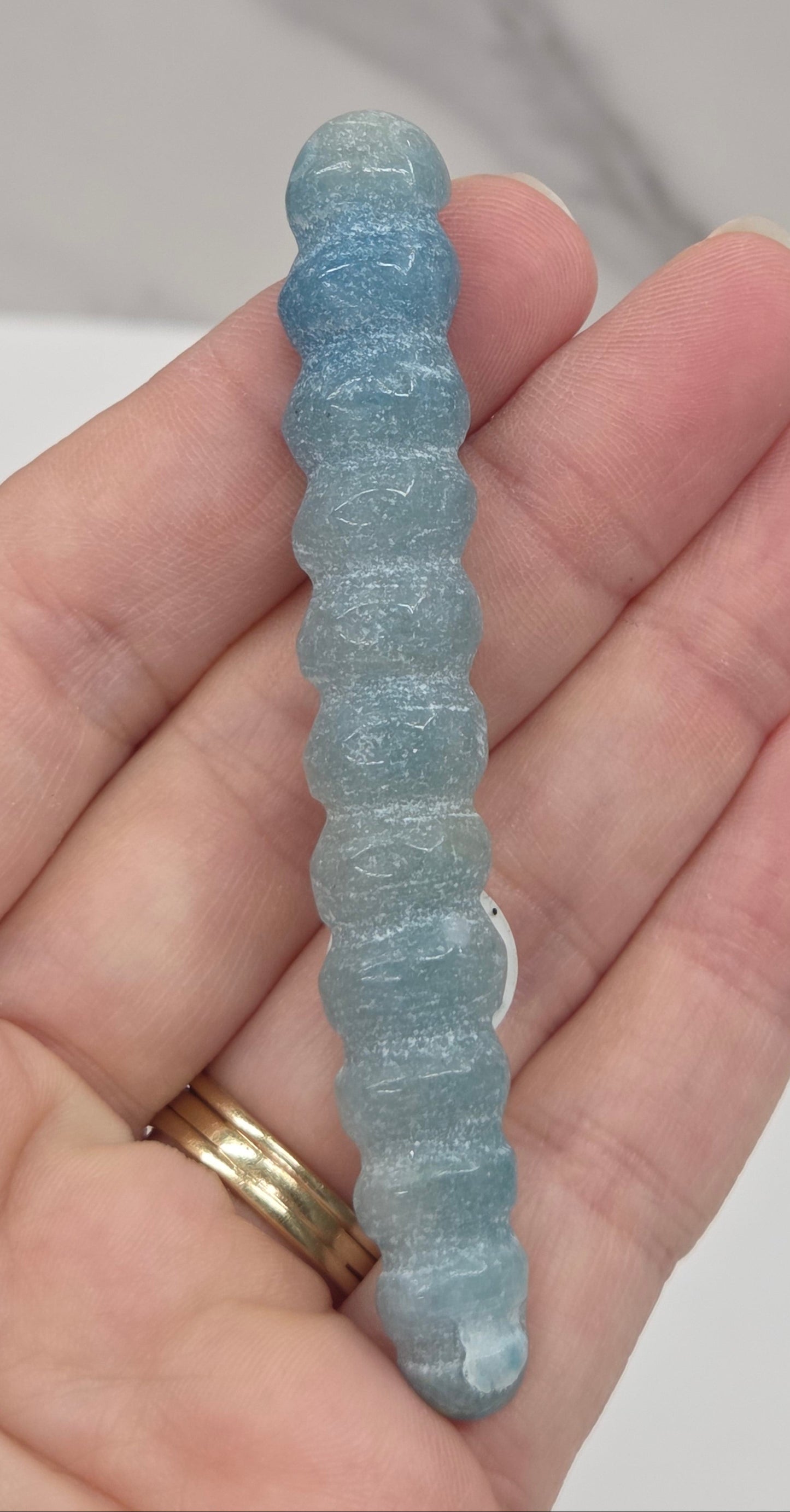 A close-up of a hand holding a light blue, ribbed, worm-like textured object, with gold rings on one finger, against a light marble-patterned background.