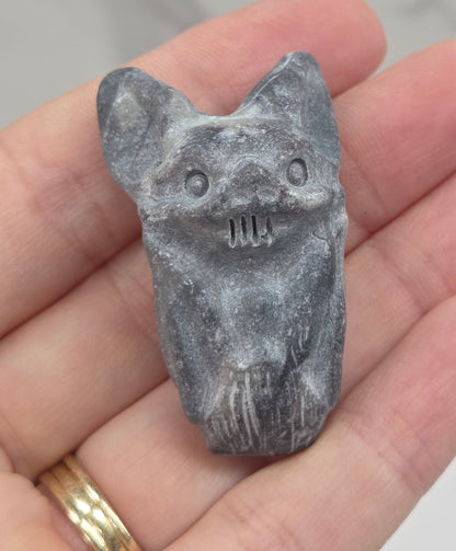 A small gray carved stone figurine shaped like a bat, held in a person's hand. The figurine has carved facial features including eyes, nose, and mouth.