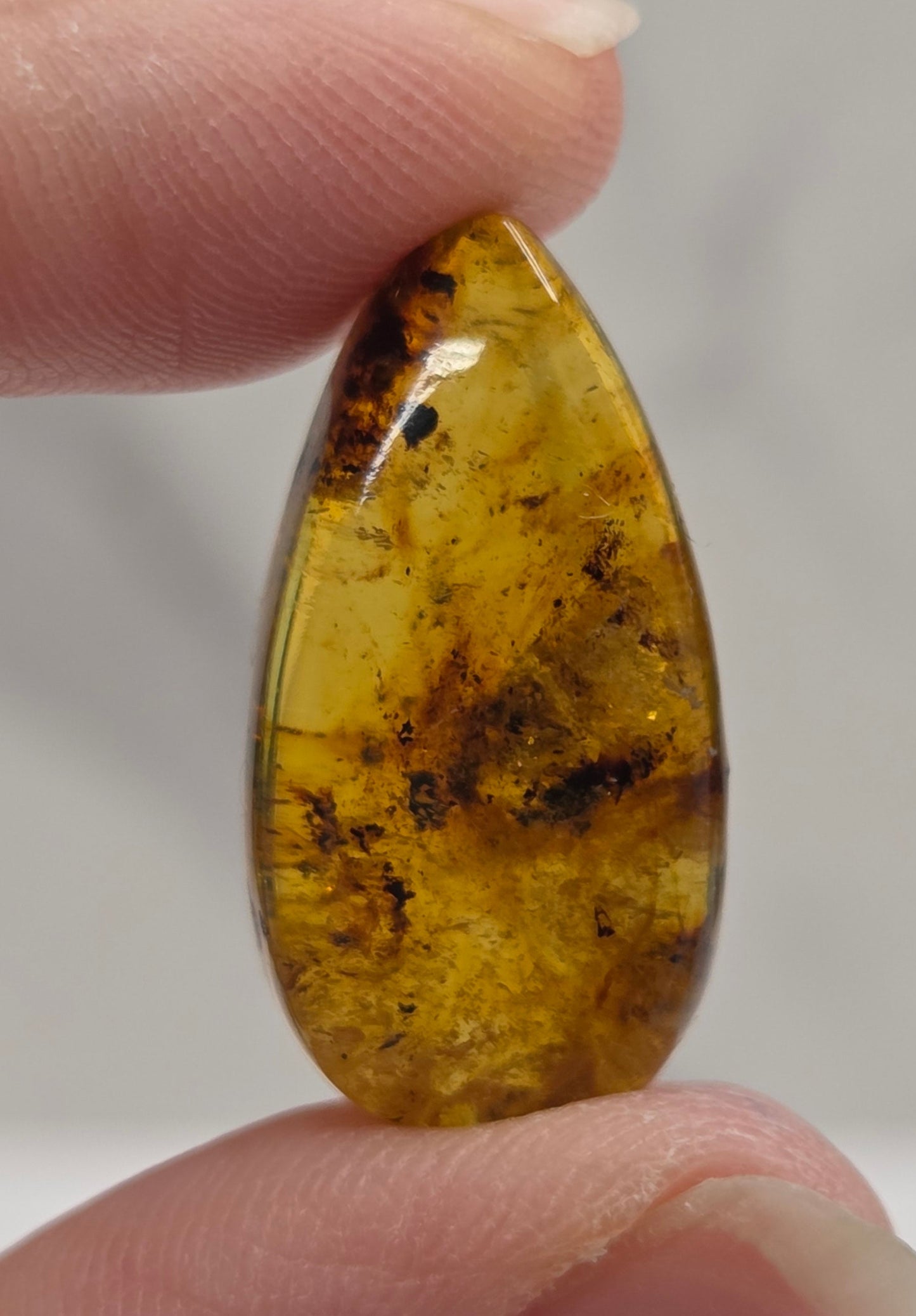 A close-up of a hand holding a polished teardrop-shaped amber gemstone with natural dark inclusions against a soft light gray background.
