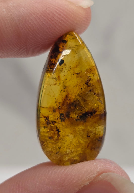 A close-up of a hand holding a polished teardrop-shaped amber gemstone with natural dark inclusions against a soft light gray background.