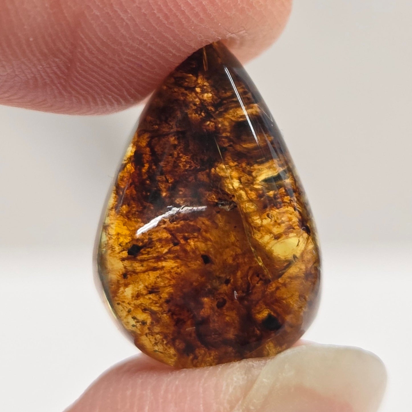 A small teardrop-shaped polished gemstone with a marbled pattern of dark brown and golden amber, held between two human fingers against a light gray background.