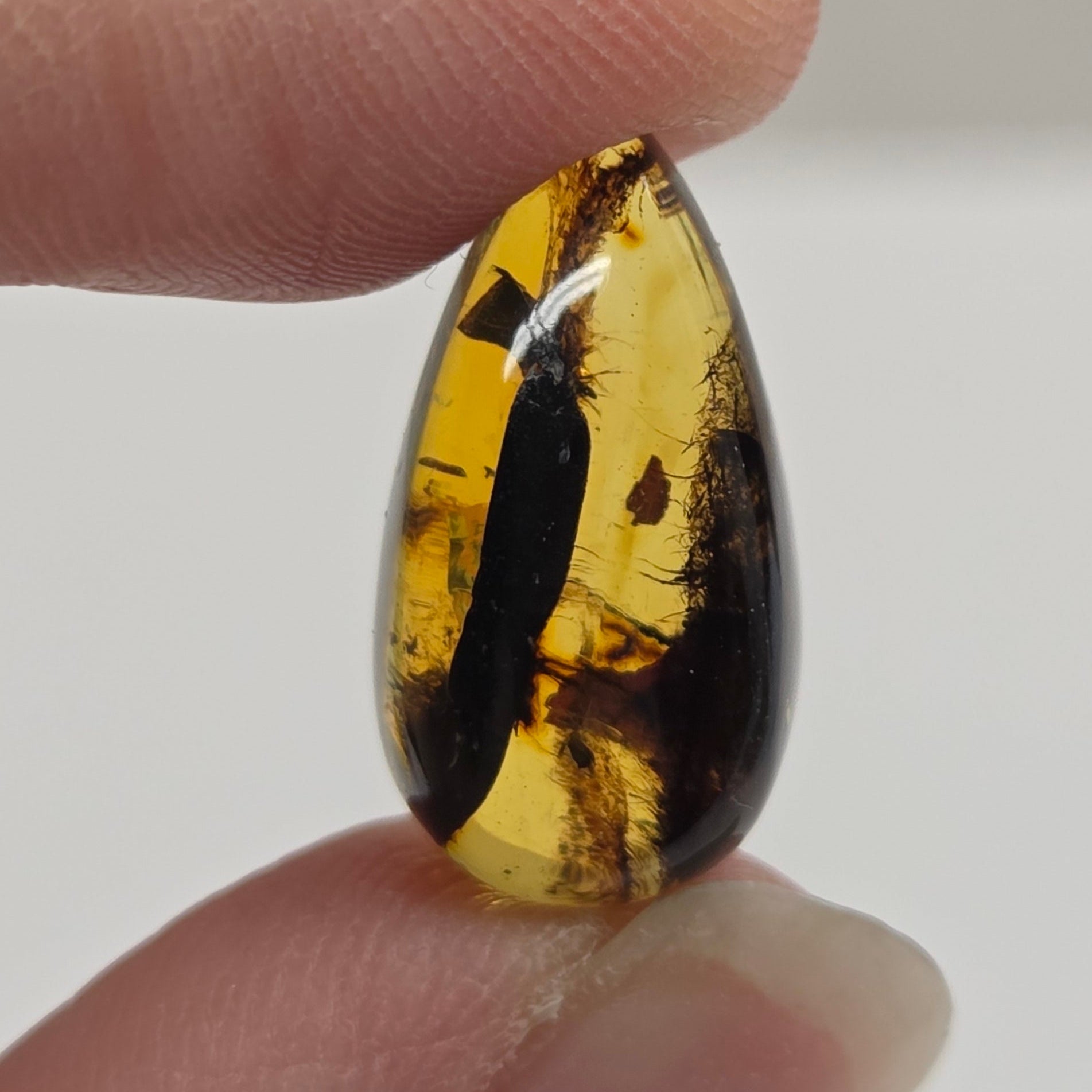 A close-up of a hand holding a polished teardrop-shaped amber gemstone with dark brown and golden yellow inclusions against a soft light gray background.