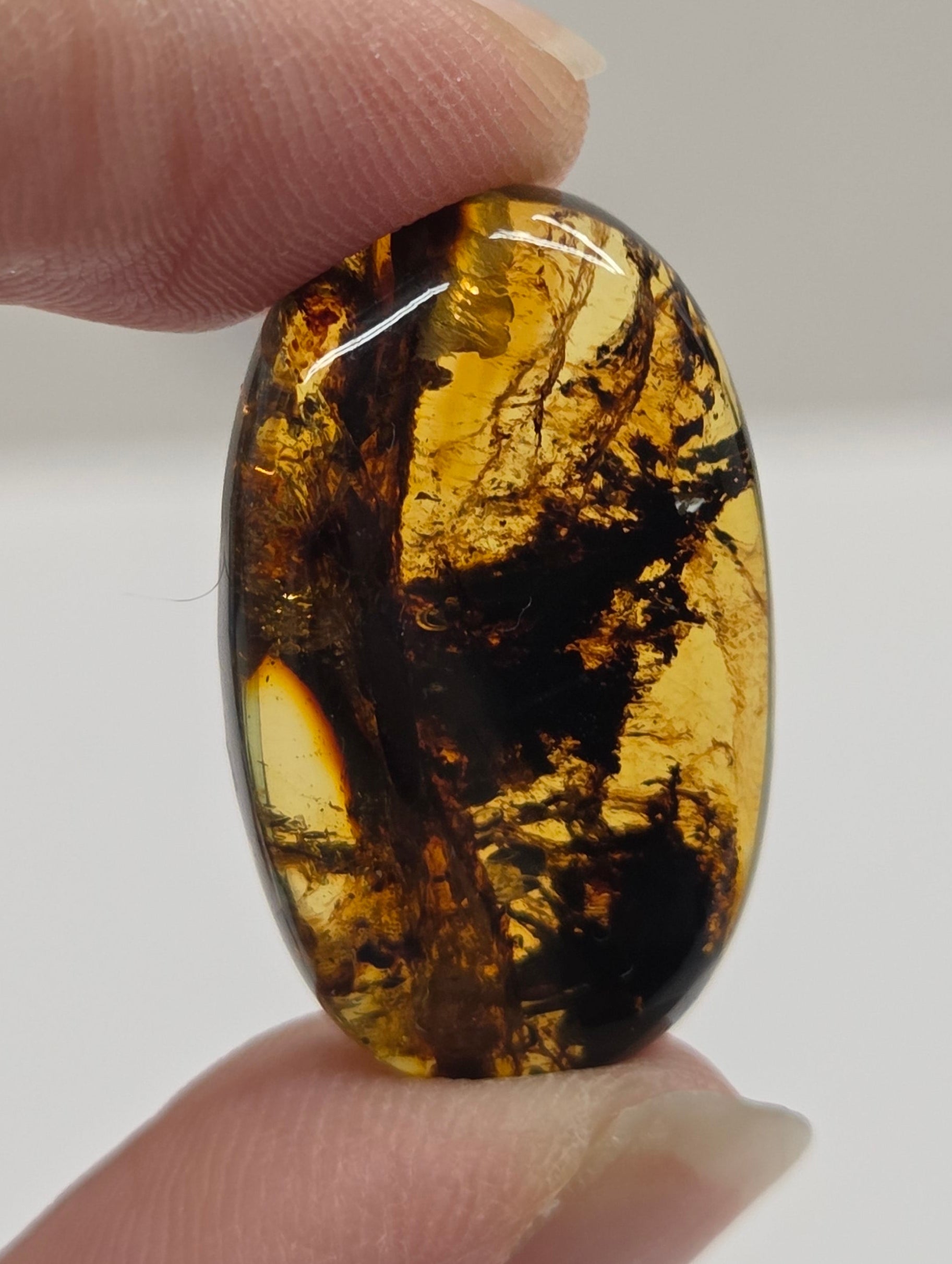 A hand holding a polished oval amber cabochon with dark inclusions, shown against a light gray background.