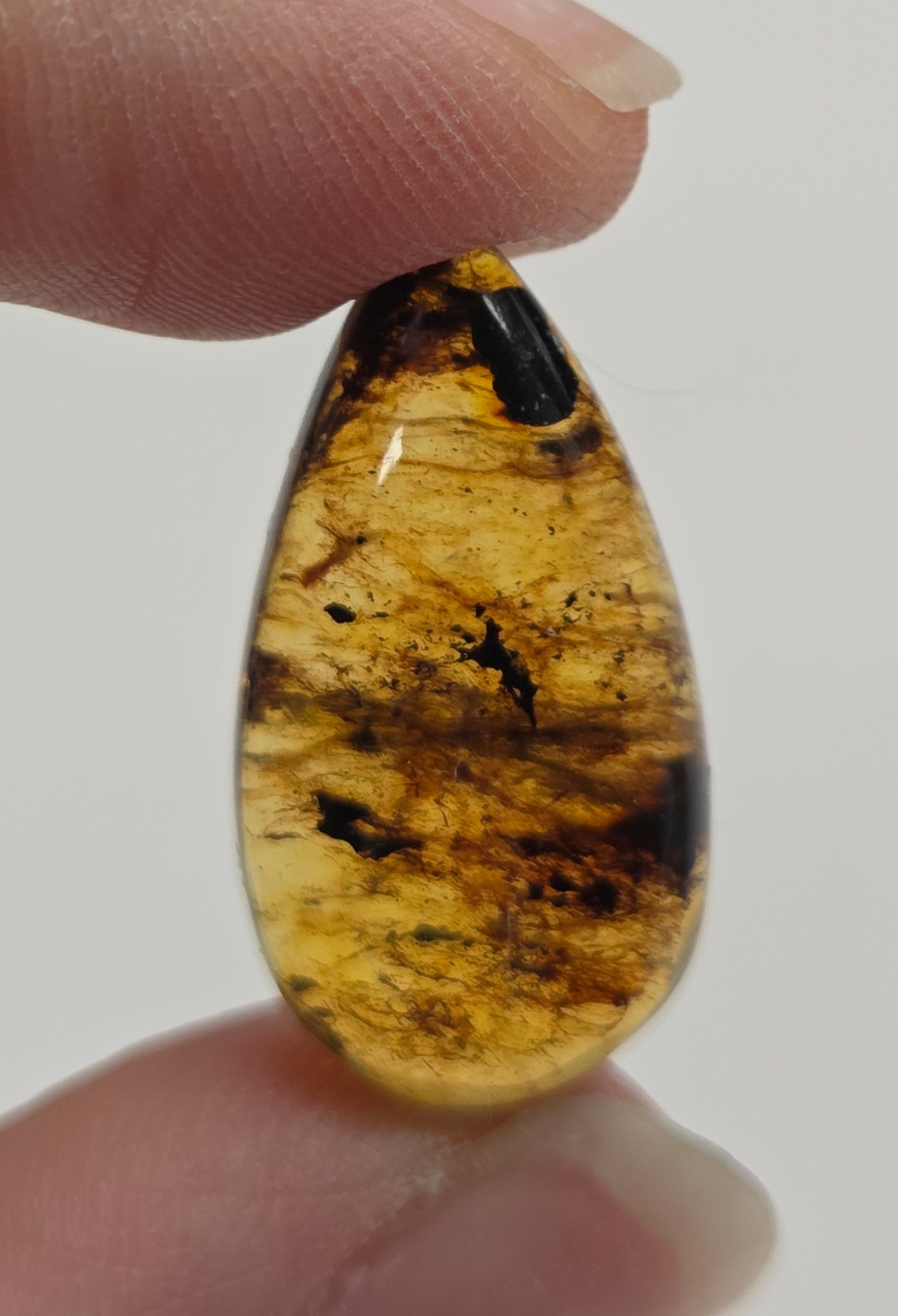A close-up of a person's fingers holding a small, teardrop-shaped cabochon gemstone with a marbled pattern of black, brown, and golden-yellow tones against a plain light gray background.