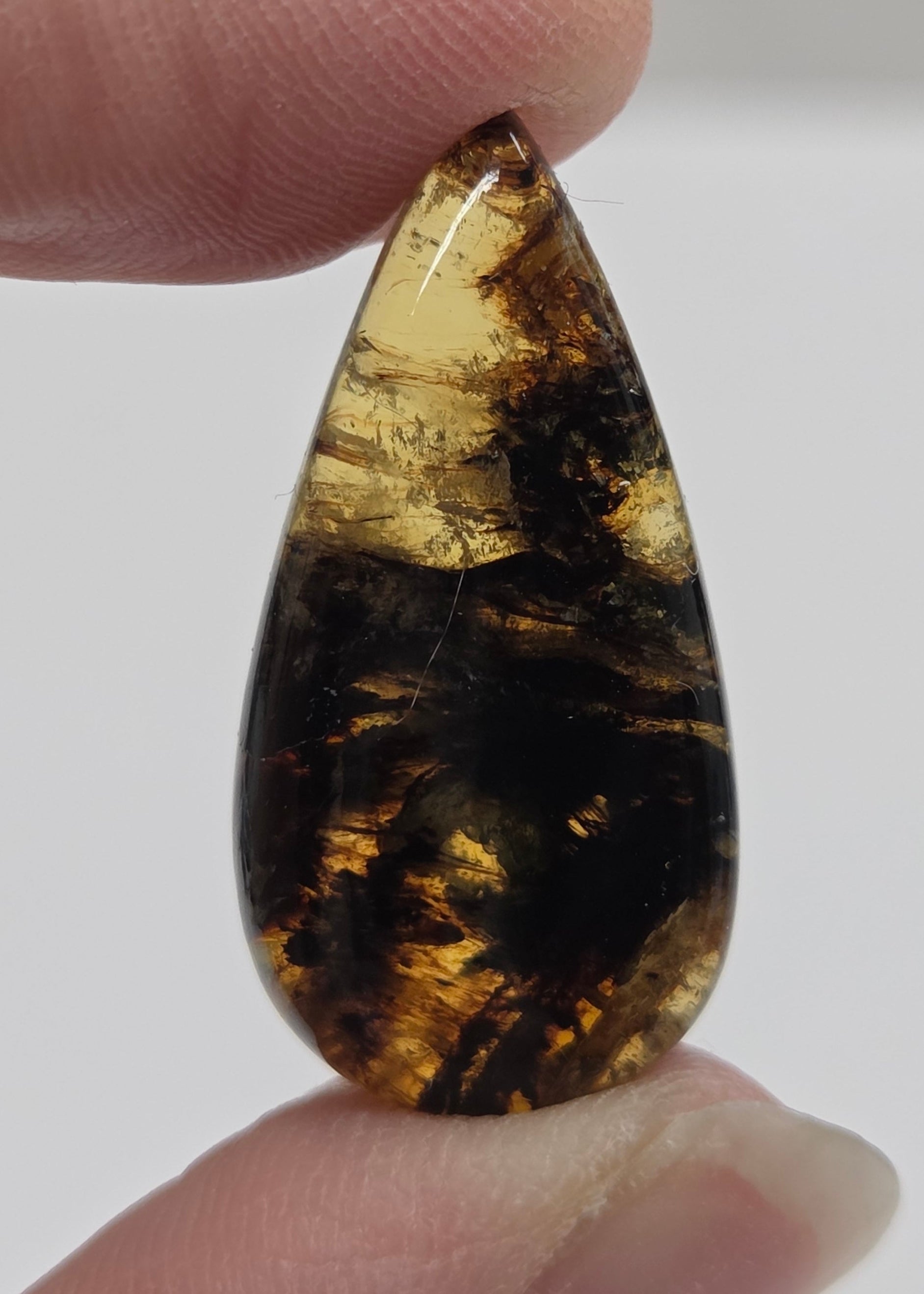 A teardrop-shaped cabochon gemstone with a dark brown and black marbled pattern and golden-brown inclusions, held between two fingers against a light gray background.