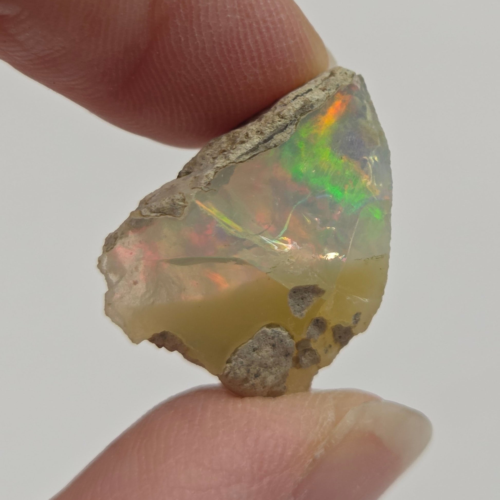A raw, rough opal gemstone held between two human fingers against a plain light gray background.