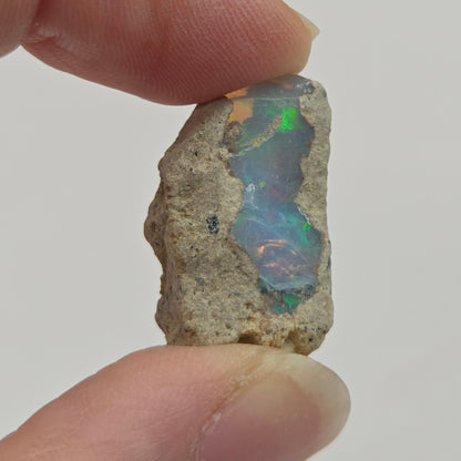 A raw opal stone held between two fingers, showing a vibrant play-of-color with blue, green, and orange hues against a neutral light gray background.