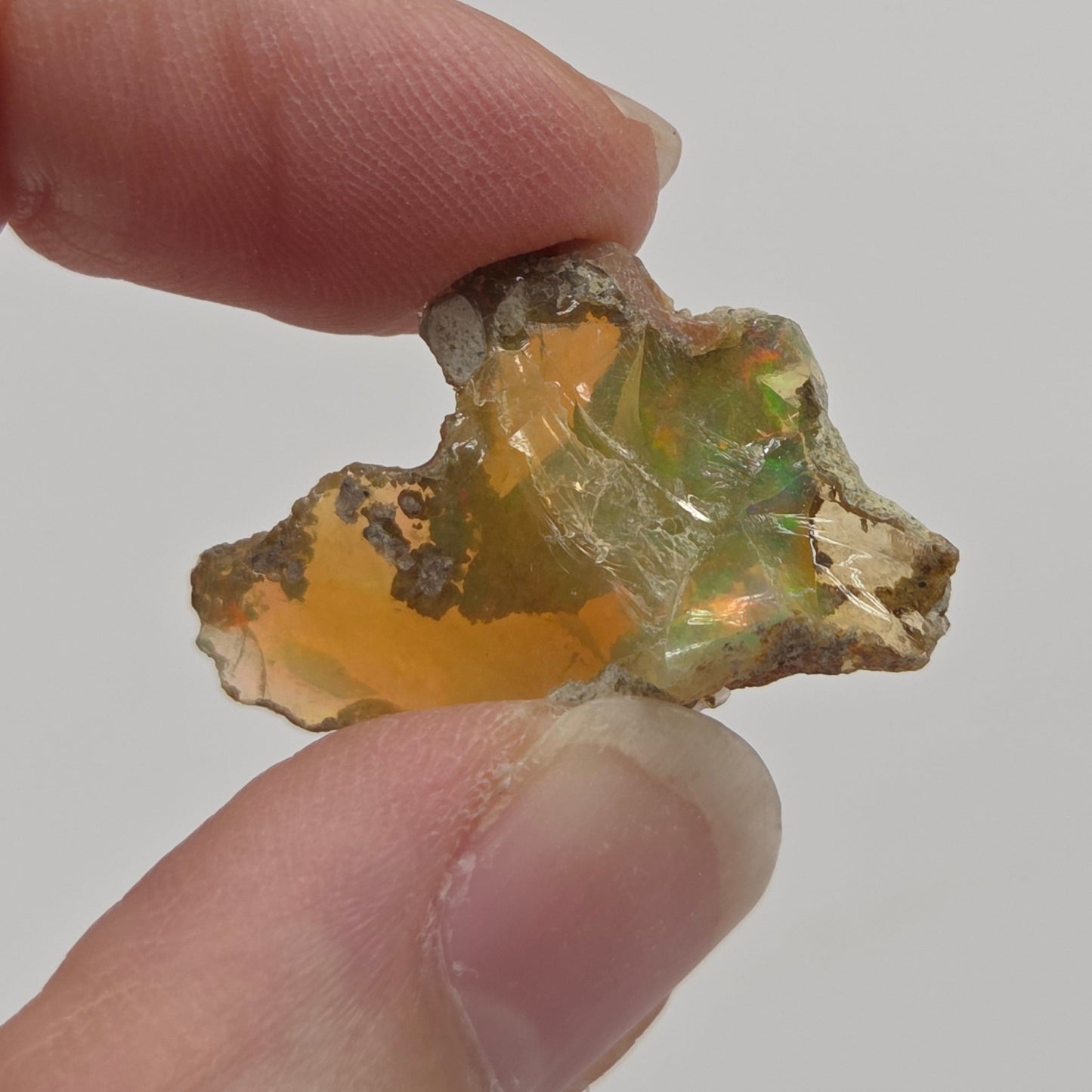 A raw, rough opalite gemstone with a golden-orange body and green flashes, held between two human fingers against a plain light gray background.