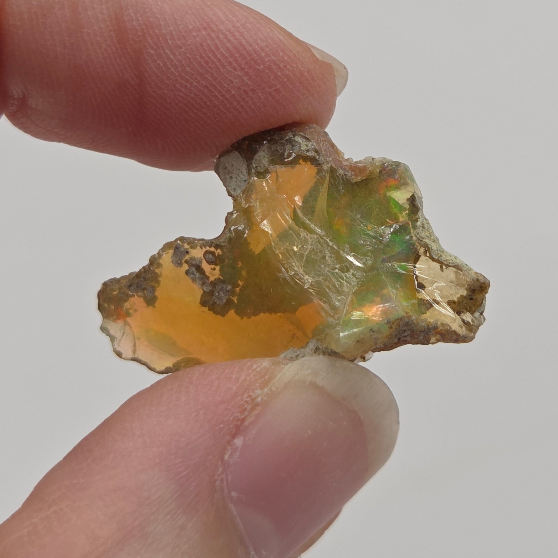 A raw, rough opalite gemstone with a golden-orange body and green flashes, held between two human fingers against a plain light gray background.