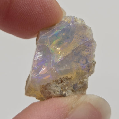 A raw, rough opalite crystal held between two human fingers to show its size and texture. The opalite displays a milky white base with vibrant, shifting iridescent flashes of blue, green, and purple.