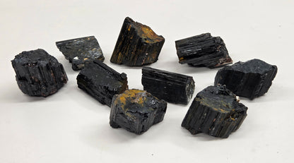 Nine rough black mineral or crystal specimens with some brownish-yellow matrix, arranged on a white surface with a light gray marble-patterned background.