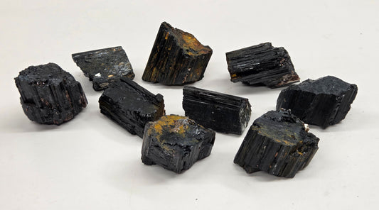 Nine rough black mineral or crystal specimens with some brownish-yellow matrix, arranged on a white surface with a light gray marble-patterned background.