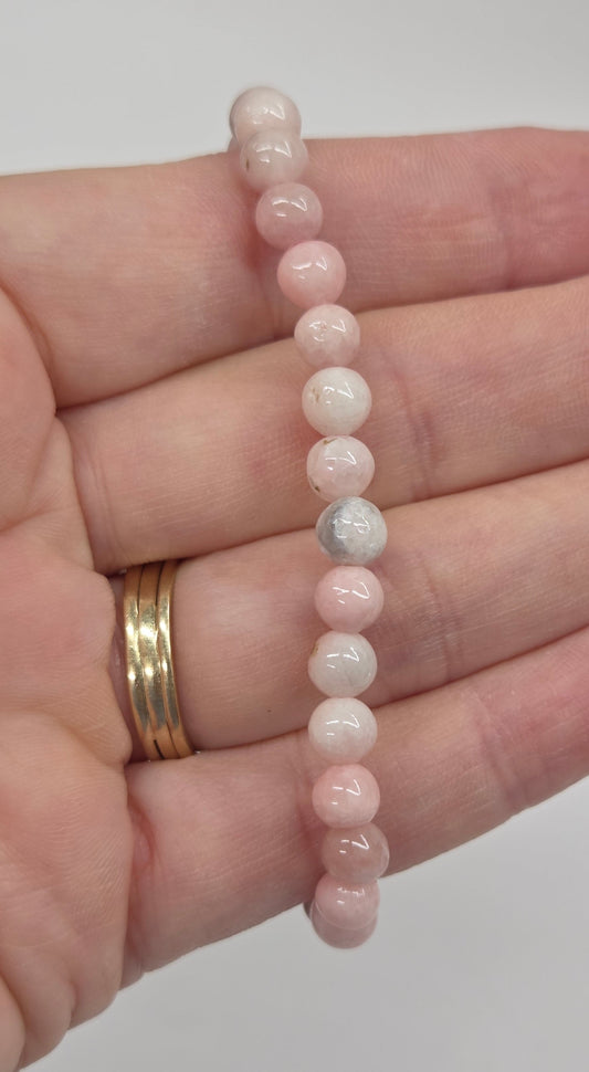 A beaded stretch bracelet made of small round pale pink and white beads, shown resting across an open hand. The hand also wears two gold rings.