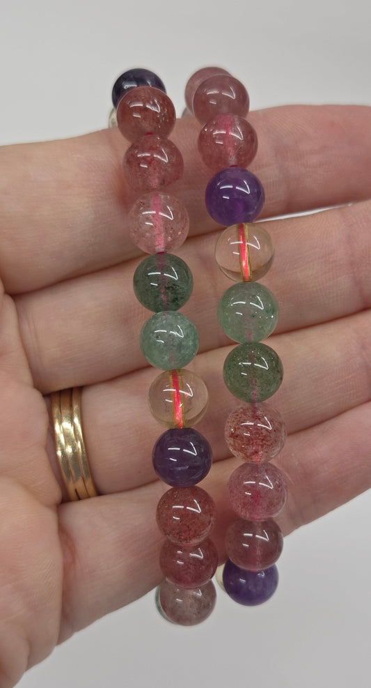 Two beaded gemstone bracelets held in a person's hand. The bracelets feature translucent round beads in pinkish-red, purple, green, and dark blue, strung on a thin red cord.