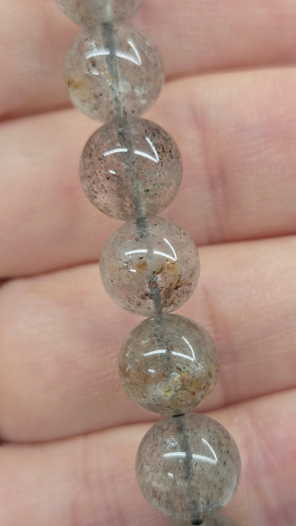 A close-up of a beaded bracelet made of translucent gray round beads with tiny dark inclusions, shown resting across a person's fingers.