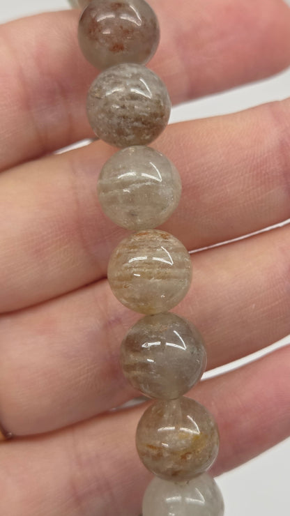 A close-up of a hand holding a strand of polished round beads made of translucent gray and brown stone, with a gold ring on one finger.