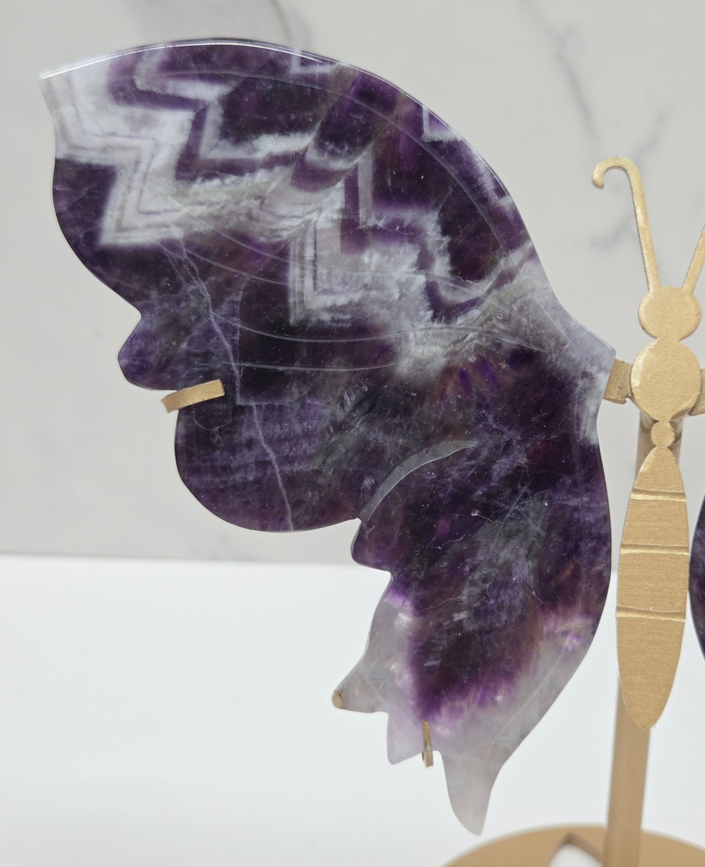 A close-up of a decorative butterfly wall art piece featuring purple and white marbled amethyst wings with a gold metal body and antennae, mounted against a white marble-patterned background.