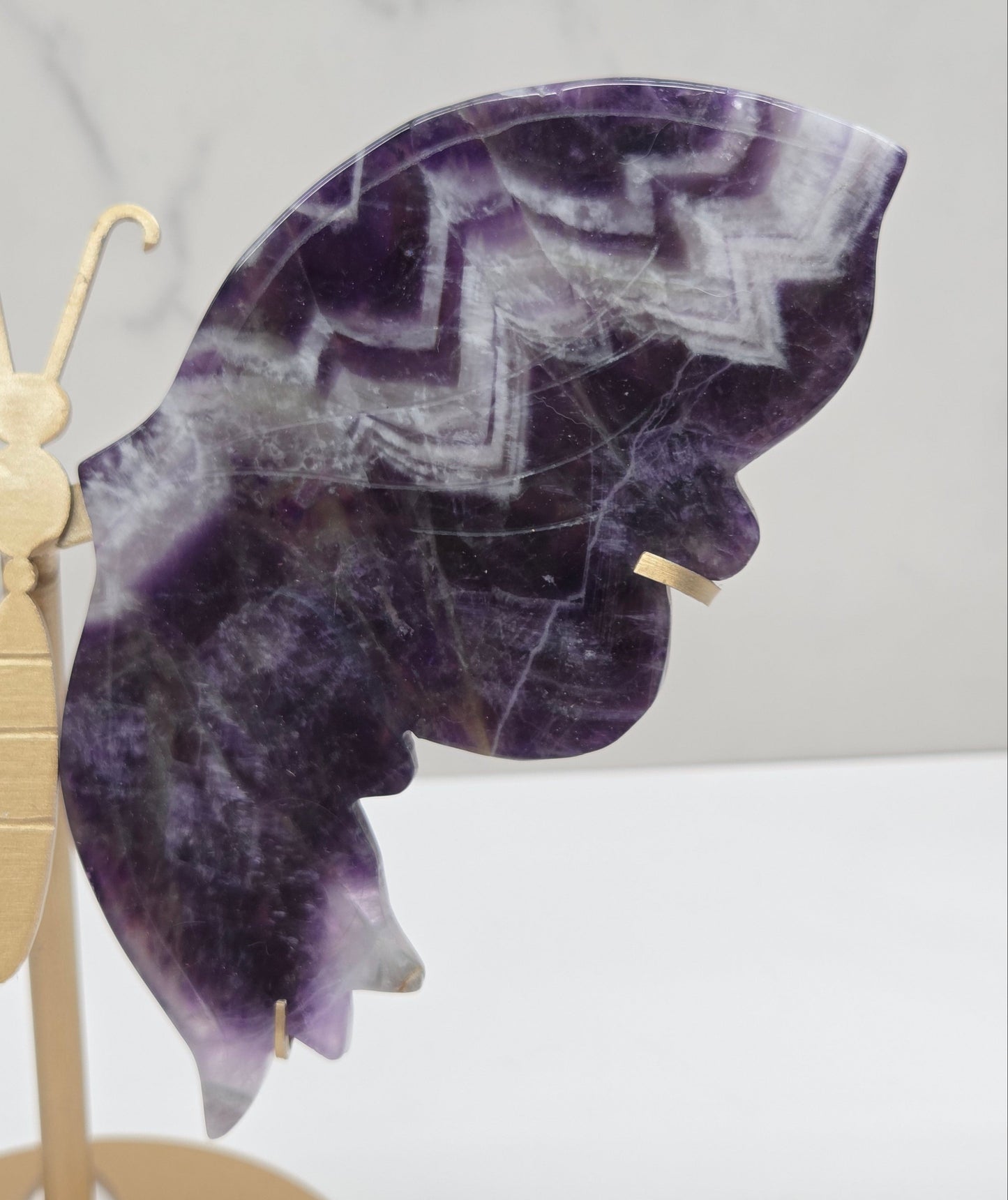 A close-up of a butterfly-shaped wall decor piece made from polished purple amethyst stone with a gold metal body and antennae, set against a light marble background.