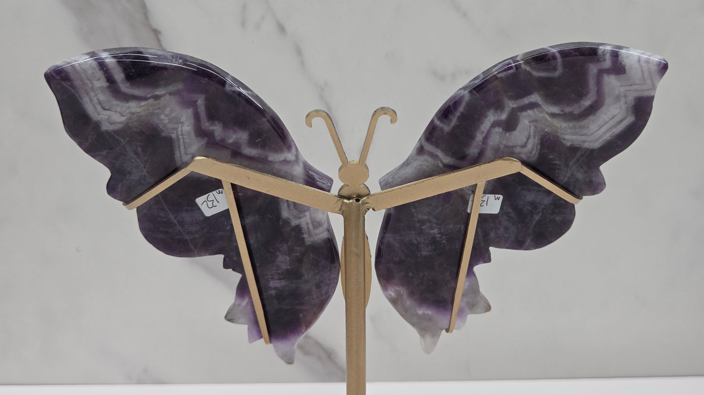 A butterfly-shaped wall sculpture made from polished purple and gray agate stone wings, mounted on a gold-toned metal stand against a white marble-patterned background.