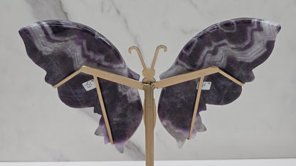 A butterfly-shaped wall sculpture made from polished purple and gray agate stone wings, mounted on a gold-toned metal stand against a white marble-patterned background.