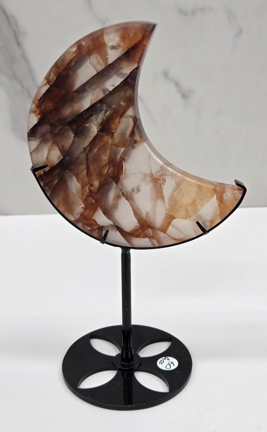 A crescent moon-shaped wall sconce with a brown and beige marbled onyx stone shade and a black metal mount, shown against a white wall with a marble-patterned surface on the left.