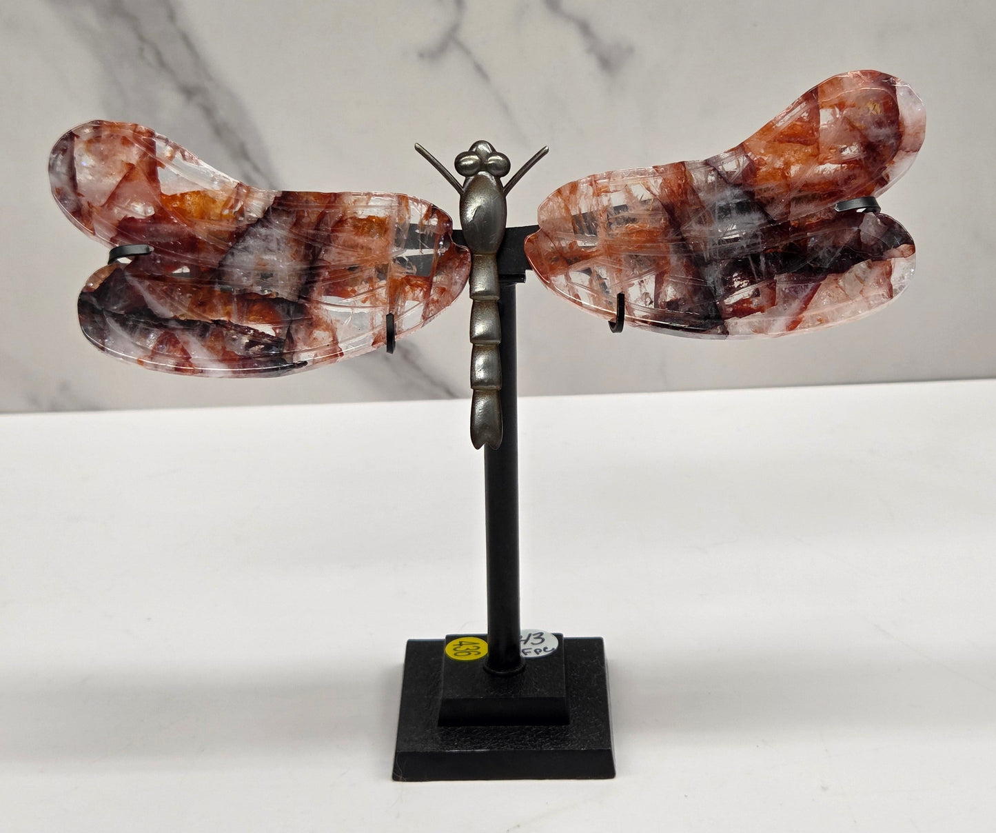 A decorative wall-mounted sculpture shaped like a butterfly or dragonfly, with marbled amber-brown and black wings attached to a black metal arm and square wall mount, shown against a white wall and light marble surface.