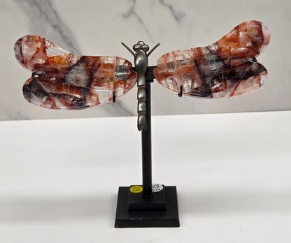 A decorative wall-mounted sculpture shaped like a butterfly or dragonfly, with marbled amber-brown and black wings attached to a black metal arm and square wall mount, shown against a white wall and light marble surface.