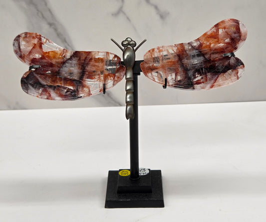 A decorative wall-mounted sculpture shaped like a butterfly or dragonfly, with marbled amber-brown and black wings attached to a black metal arm and square wall mount, shown against a white wall and light marble surface.