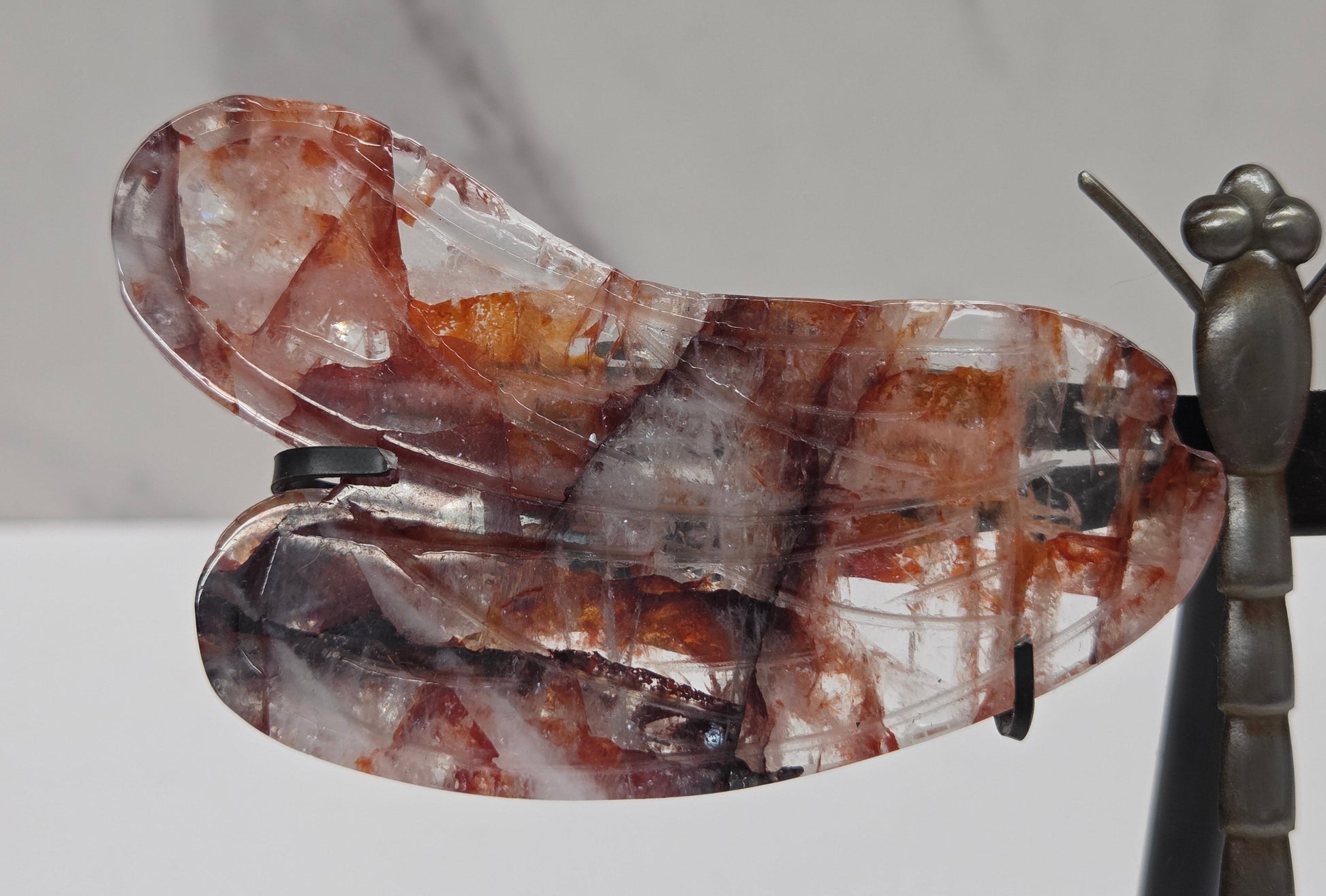 A dragonfly wing pendant made of clear quartz with brown and amber inclusions, displayed on a black stand next to a metallic dragonfly figurine against a white marble background.