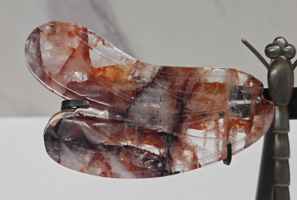 A dragonfly wing pendant made of clear quartz with brown and amber inclusions, displayed on a black stand next to a metallic dragonfly figurine against a white marble background.