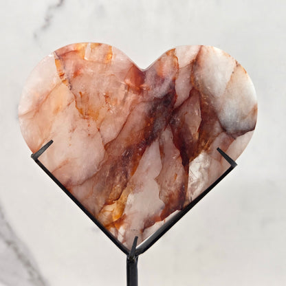 Fire Quartz heart on a black stand, shown against a white wall and marble-patterned background.