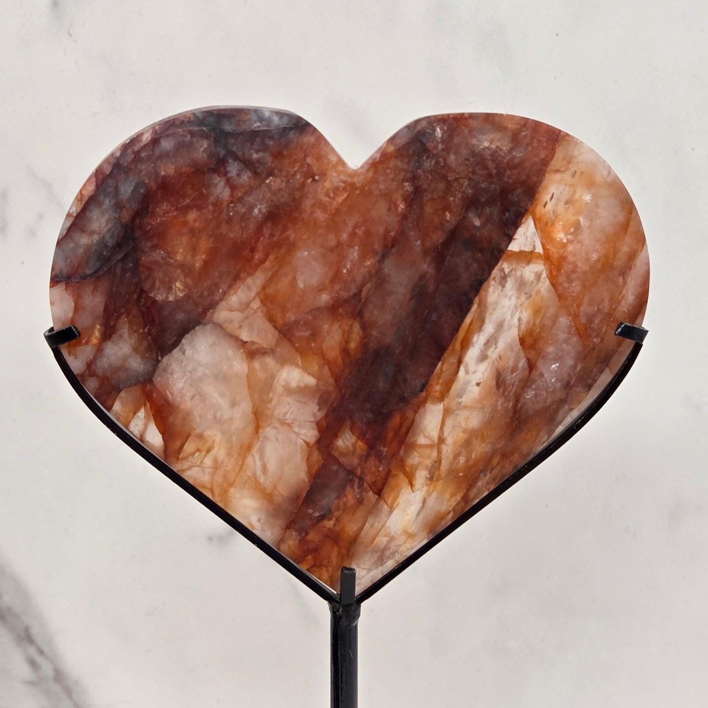 Fire Quartz heart on a black stand, shown against a white wall and marble-patterned background.