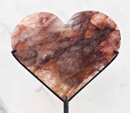 Fire Quartz heart on a black stand, shown against a white wall and marble-patterned background.