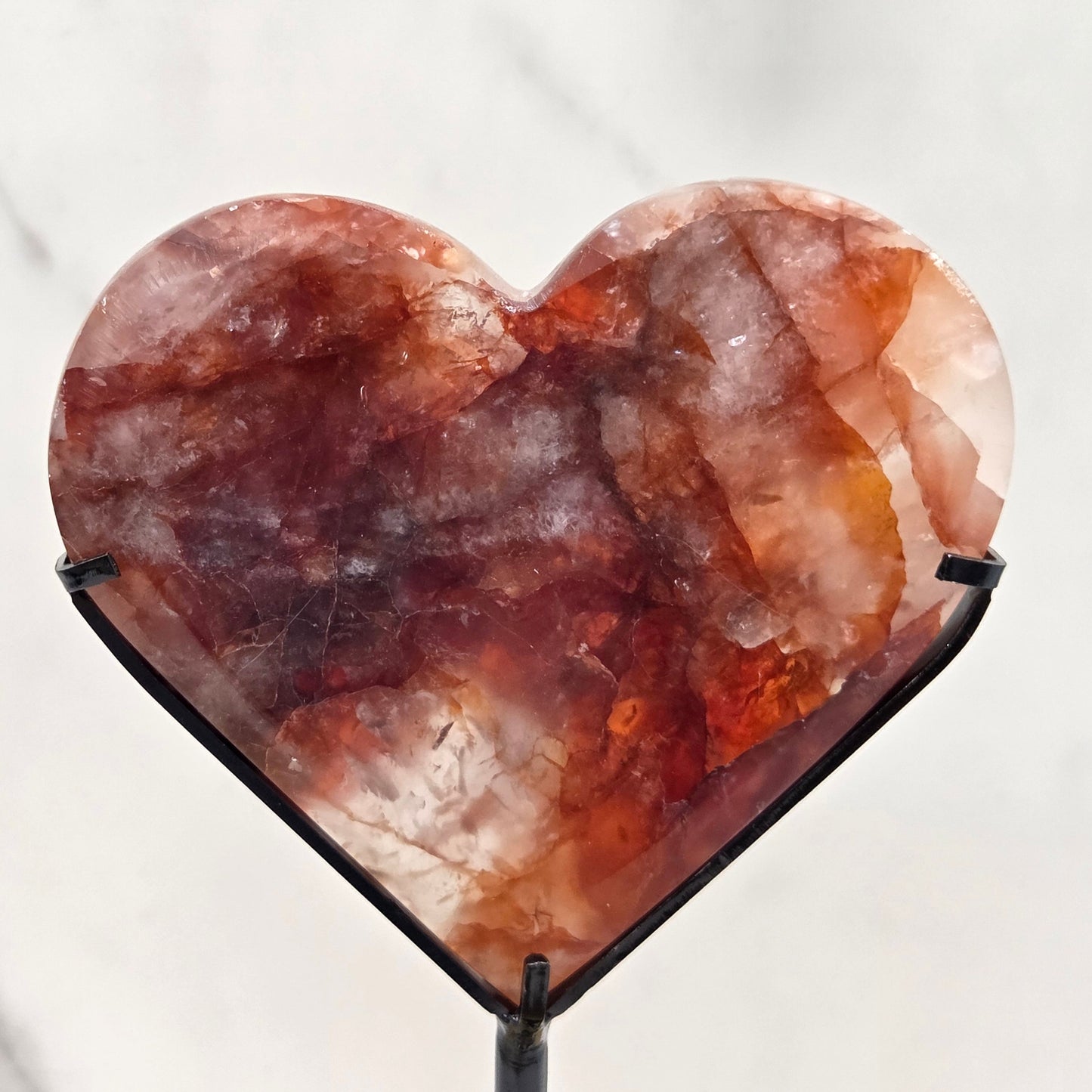 Fire Quartz heart on a black stand, shown against a white wall and marble-patterned background.