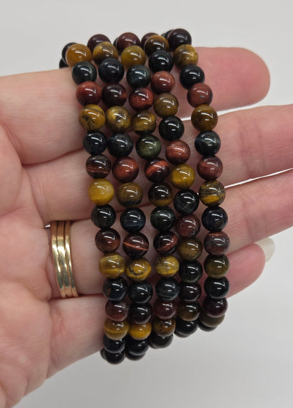 A hand holding three beaded bracelets made of polished round stones in black, brown, and golden-yellow tiger-eye patterns, with a gold ring on the finger, against a plain light gray background.