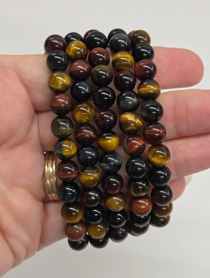 A hand holding a stack of five beaded stretch bracelets made of polished round stones with black, brown, and golden-yellow tiger-eye patterns, against a plain white background.