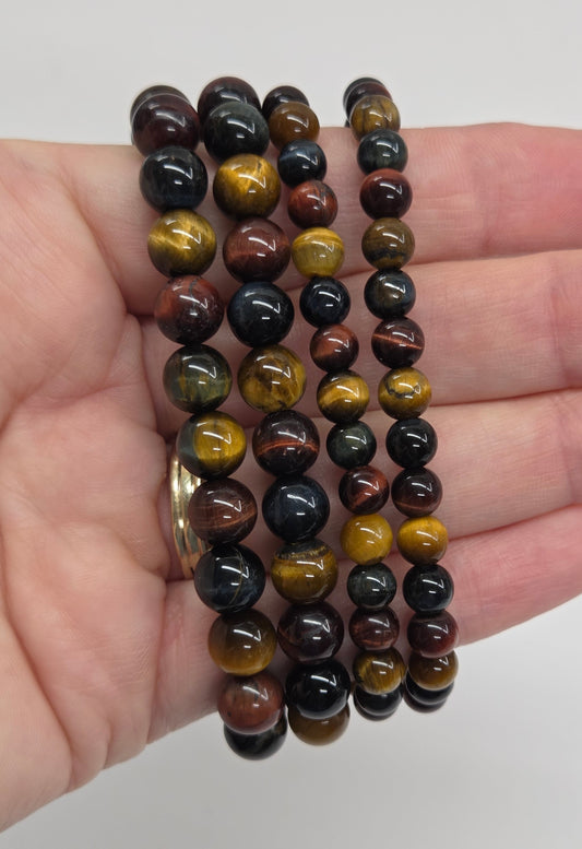 A hand holding four beaded stretch bracelets made of polished round beads in black, brown, and golden yellow tiger's eye pattern, against a plain light background.