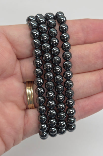 A hand holding three rows of small dark metallic magnetic beads, shown against a plain light background. A gold ring is visible on the finger for scale.