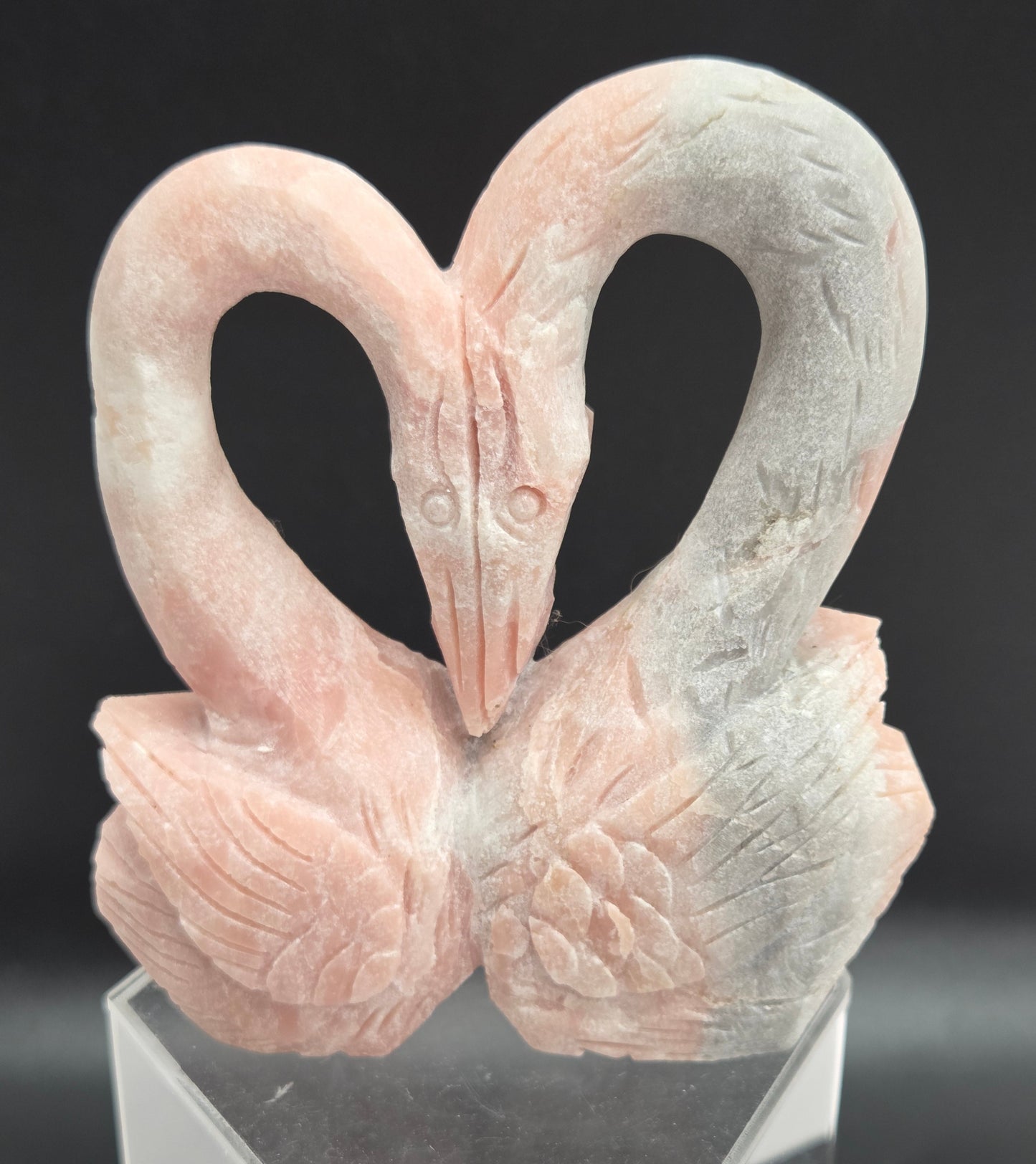 A carved decorative object shaped like two intertwined swans or birds, shown close up against a dark background. The item appears pale pink to off-white with a smooth, polished finish and is mounted on a clear acrylic or glass stand.